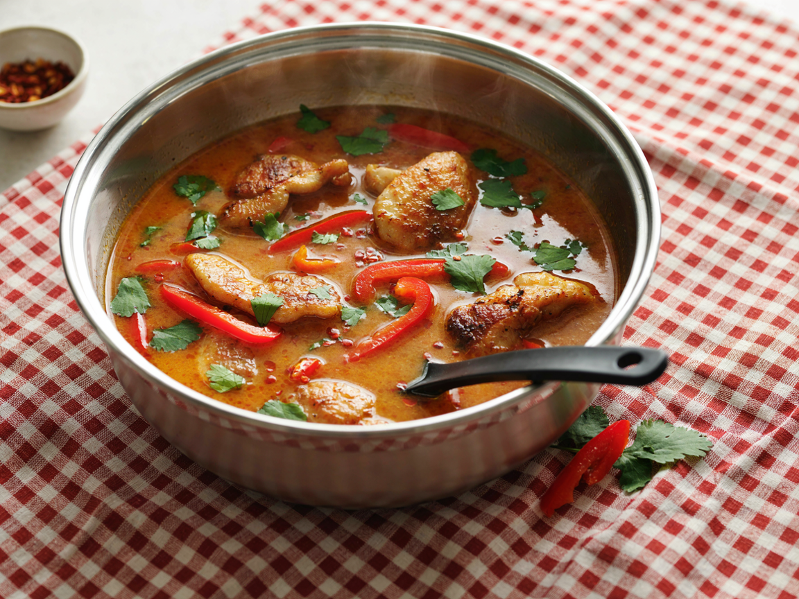 A wide stainless steel pot of creamy Thai red curry seen from above at an angle, golden-seared chicken pieces and soft red bell pepper strips in a glossy orange-red coconut sauce with beads of red oil, scattered fresh cilantro, a black serving spoon resting in the pot, on a red and white gingham cloth, warm natural light, food photography