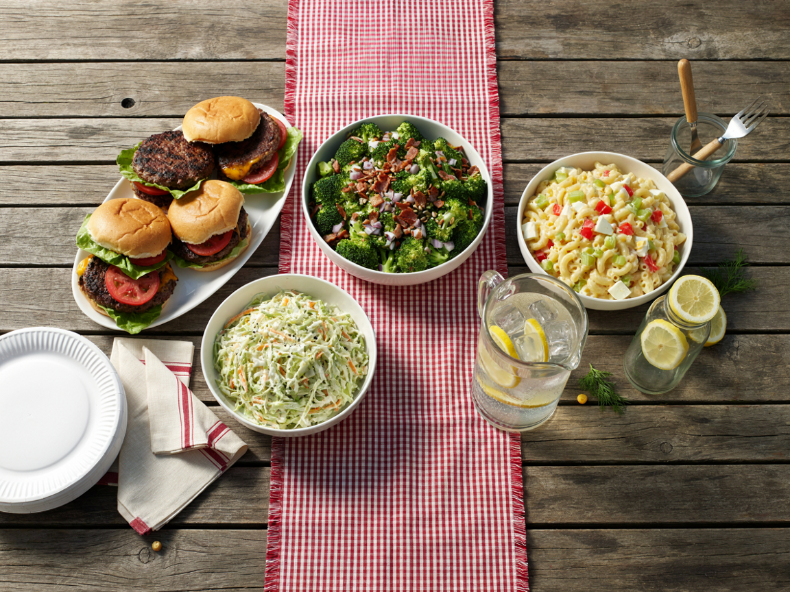 Cookout table with a large bowl of broccoli salad beside burgers, macaroni salad, coleslaw and lemonade