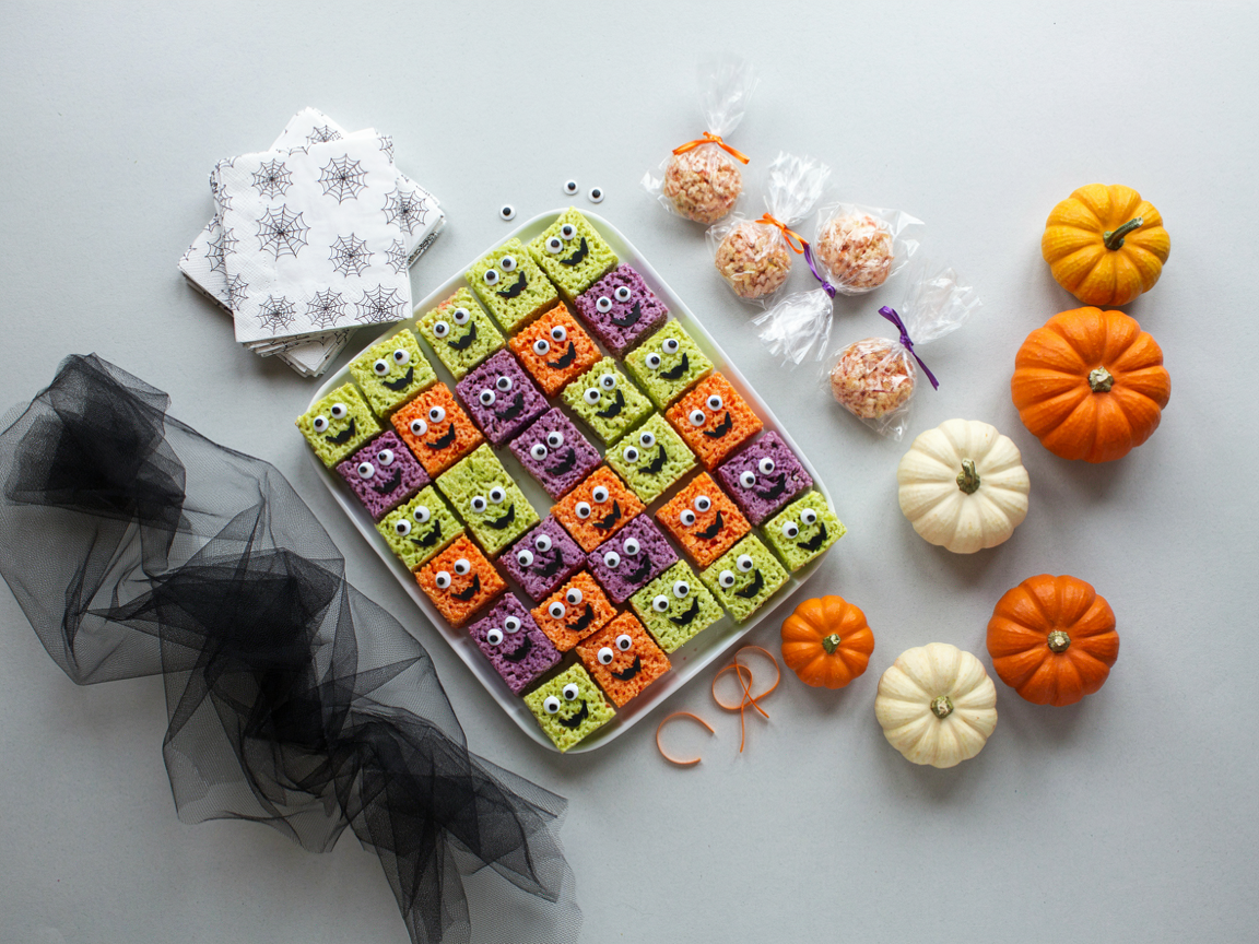 A Halloween class party table seen from above: a tray of monster crispy rice cereal treats in lime green, purple and orange with candy eyes, some wrapped in clear cellophane bags with orange and purple twist ties, small paper napkins, mini pumpkins and black tulle on a light grey table, bright daylight