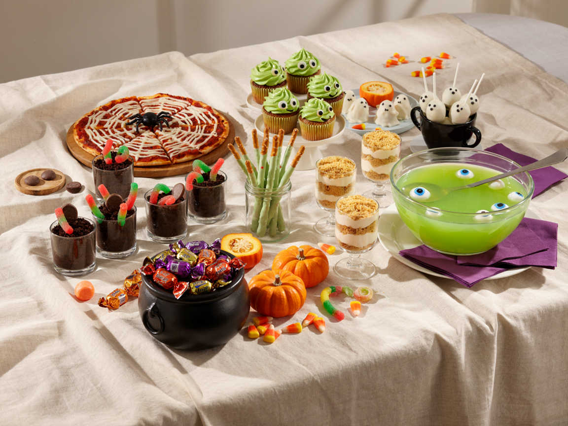 A Halloween party food table seen from above at an angle on a cream tablecloth with orange and purple accents: a spider web pizza on a wooden board, green monster cupcakes with candy eyes, ghost cake pops standing in a black cup, chocolate dirt cups with gummy worms and cookie tombstones, green witch-finger pretzel rods in a jar, candy corn layered parfait cups, a glass punch bowl of bright green punch with a ladle, small pumpkins, warm light