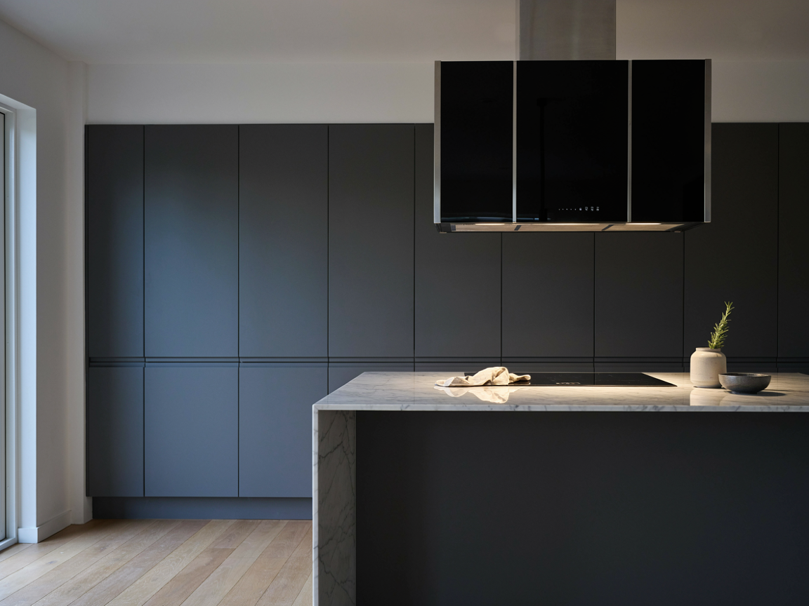 Minimalist kitchen with a black glass wall mount range hood above an induction cooktop and matte charcoal cabinets