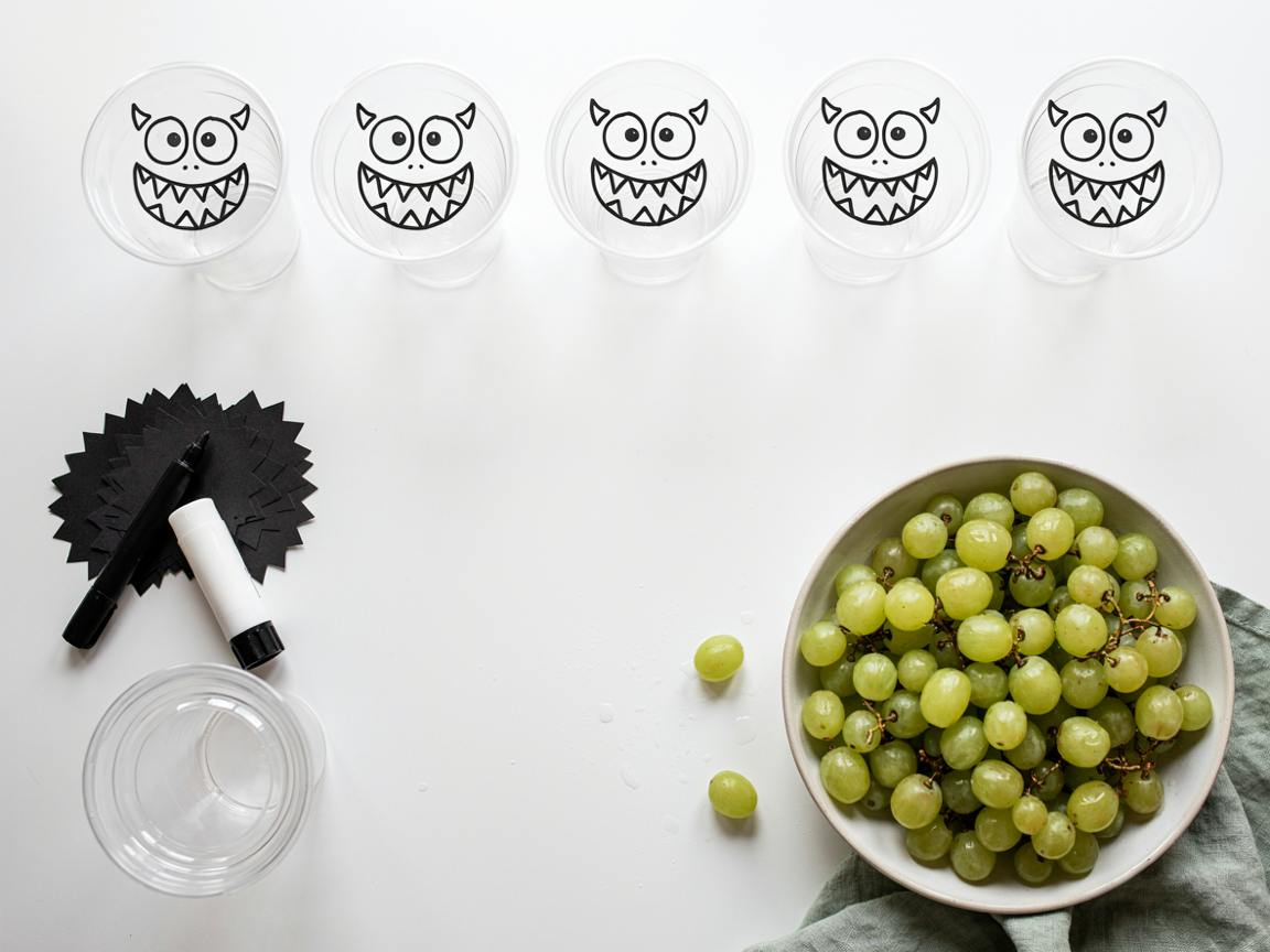 A craft and snack assembly line on a white table from above: a row of empty clear plastic cups with cartoon monster faces drawn in black marker, a stack of black paper strips cut with a zigzag edge, a black permanent marker, a glue stick, a large bowl of washed green grapes on a kitchen towel and a stack of clear lids, bright even light