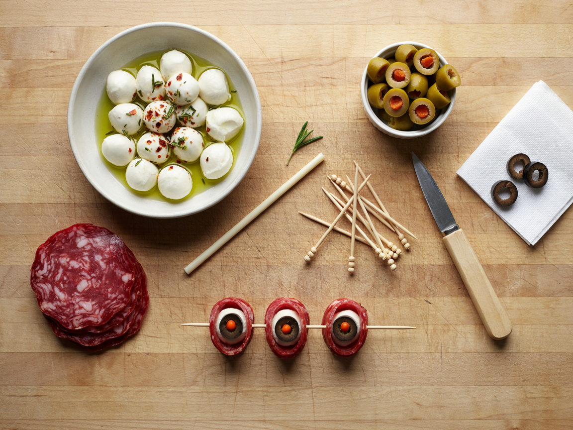 Assembling mozzarella eyeball skewers on a wooden board seen from above: a bowl of mini mozzarella balls glossy with olive oil, chili flakes and chopped rosemary, a dish of pimento-stuffed green olives, black olive rings drying on paper towel, a stack of folded red salami slices, bamboo knot picks, a drinking straw and a small paring knife, a few finished skewers in a row, soft daylight
