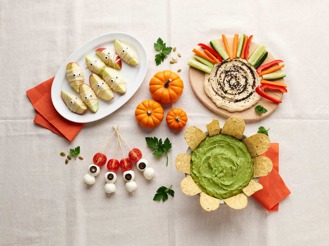A Halloween snack table for kids seen from above on a cream tablecloth: a platter of green and red mummy apple bites with white chocolate bandages and candy eyes, a round board of hummus with a spider web pattern and vegetable sticks, eyeball caprese skewers of mozzarella balls with olive pupils and cherry tomatoes on party picks, a dish of guacamole with tortilla chip tombstones, small pumpkins and orange napkins, bright daylight