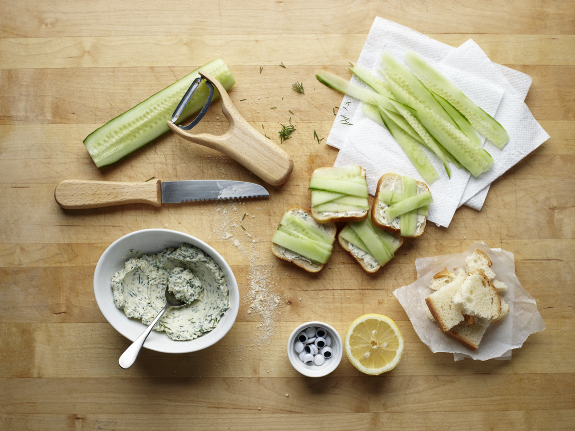 Making mummy cucumber tea sandwiches on a light wooden board seen from above: an English cucumber with a Y-shaped vegetable peeler, long thin pale-green ribbons drying on paper towels, a bowl of herbed cream cheese flecked with dill and chives, crustless white bread rectangles spread with filling, a serrated knife, trimmed crusts, a small dish of candy eyes and a lemon