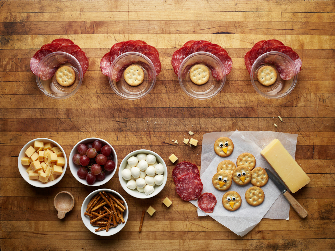 Assembling turkey charcuterie cups on a wooden board, seen from above: a row of clear cups with thin salami slices fanned around the back of each, round crackers with candy eyes and cheddar beaks laid out to set, small bowls of cheddar cubes, red grapes, mini mozzarella balls and pretzel sticks, a small knife and a block of cheddar, warm daylight