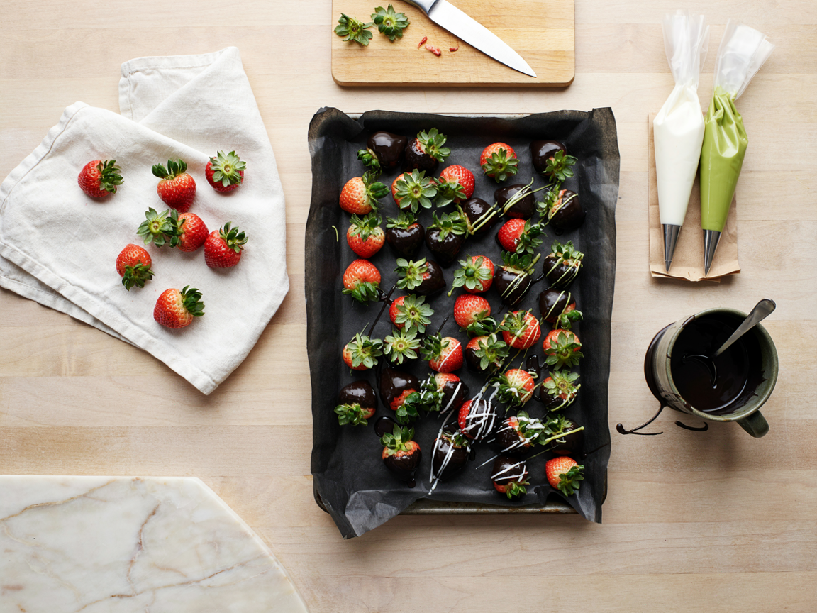 Dipping strawberries on a kitchen counter seen from above: a parchment-lined sheet pan with rows of strawberries freshly dipped in glossy black coating, a deep mug of melted black coating, two small piping bags of white and lime-green coating with snipped tips, washed strawberries drying on a white towel, soft daylight