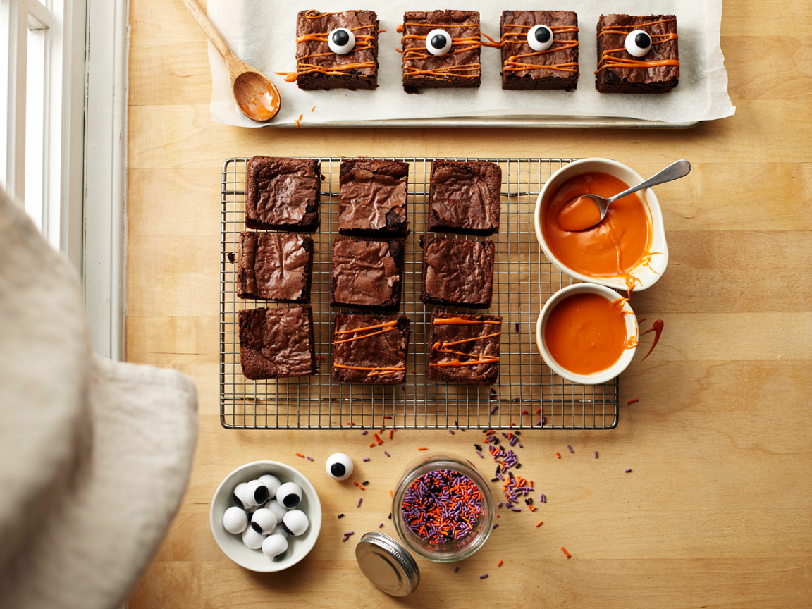 A kitchen counter from above: a wire rack of cooled fudgy brownie squares, a small bowl of melted orange candy melts with a spoon drizzling zigzags over the squares, a dish of candy eyeballs in three sizes, a jar of orange, purple and black sprinkles, a parchment-lined tray with a few finished eyeball brownies, warm kitchen light