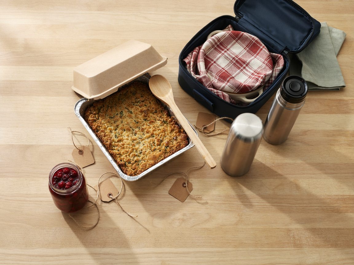 Thanksgiving side dishes packed to travel: foil pans with lids, a casserole dish wrapped in a towel inside a carrier, a thermos and a serving spoon