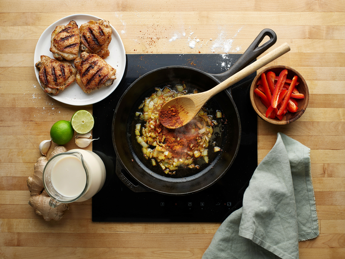 A large deep skillet on a stovetop from above, softened golden onions with curry powder, turmeric and tomato paste darkening in oil, a wooden spoon resting in the pan, a plate of seared chicken pieces, a bowl of sliced red bell pepper strips, an open glass jug of coconut milk, garlic, ginger and a lime on the counter beside it, warm kitchen light