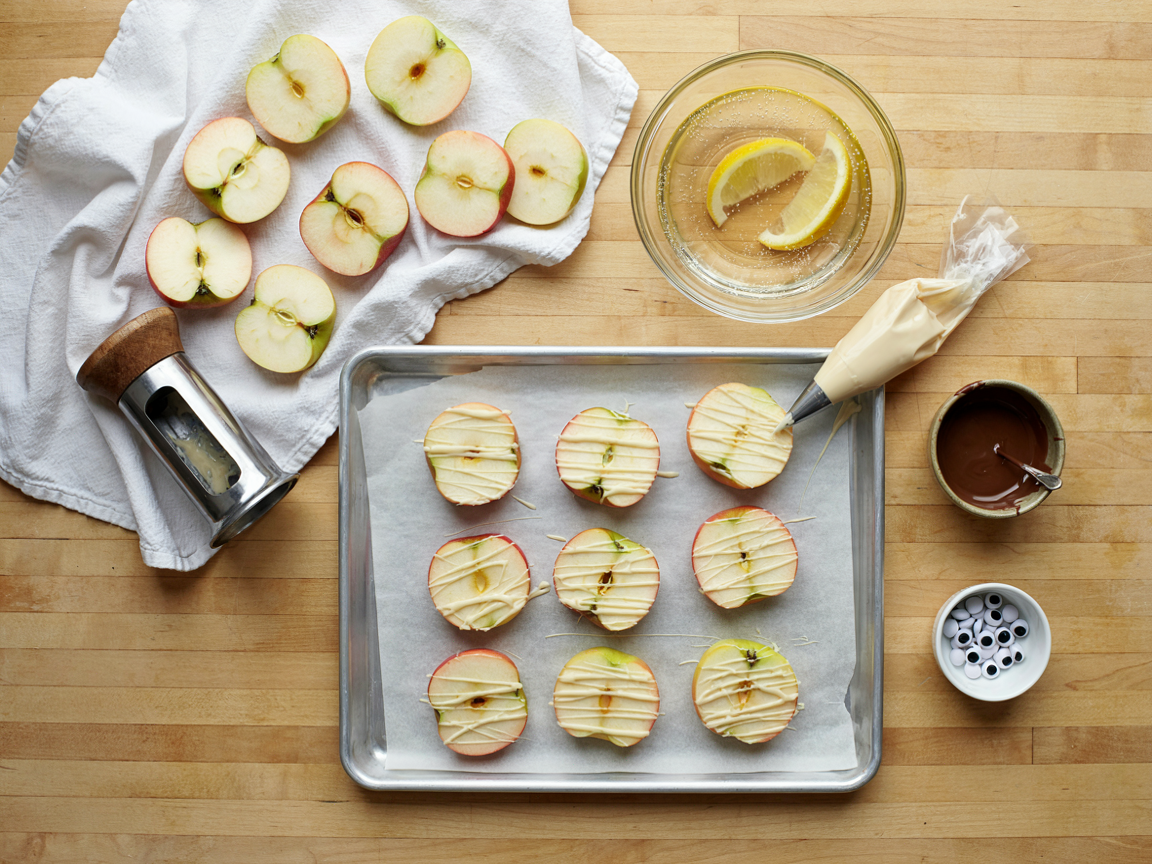 Making mummy apple bites on a light wooden counter, seen from above: cored green and red apple rounds drying on a clean kitchen towel, a glass bowl of lemon water, an apple corer, a parchment-lined tray of rounds half covered in white chocolate zigzags, a small piping bag of melted white chocolate, a bowl of melted dark chocolate and a dish of candy eyes, bright daylight