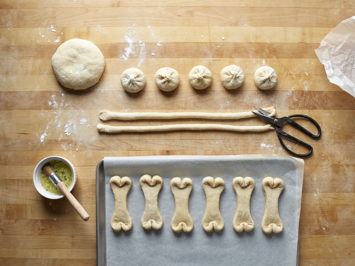 Shaping breadstick bones on a lightly floured wooden counter seen from above: a ball of pizza dough, small dough pieces, rolled dough ropes, kitchen scissors beside ropes with snipped ends, finished bone shapes with curled knobs on a parchment-lined baking sheet, a small bowl of garlic butter with a pastry brush