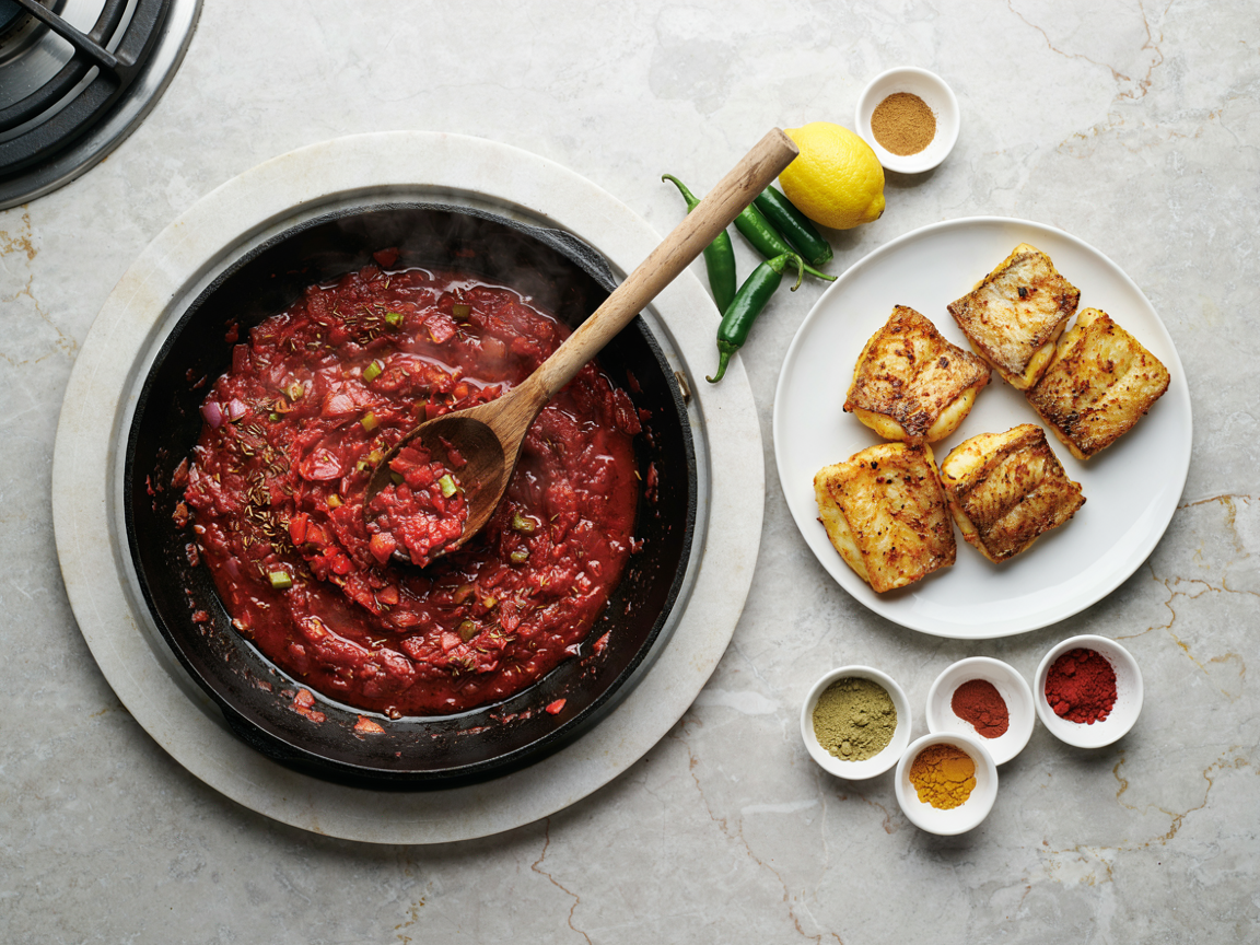 A wide pan on a stovetop from above with thick dark-red tomato and onion masala cooked down until oil glistens at the edges, a wooden spoon resting in it, a plate of golden seared turmeric-marinated fish chunks beside the pan, small bowls of ground coriander, cumin, Kashmiri chilli and turmeric, green chillies and a lemon on the counter, warm kitchen light