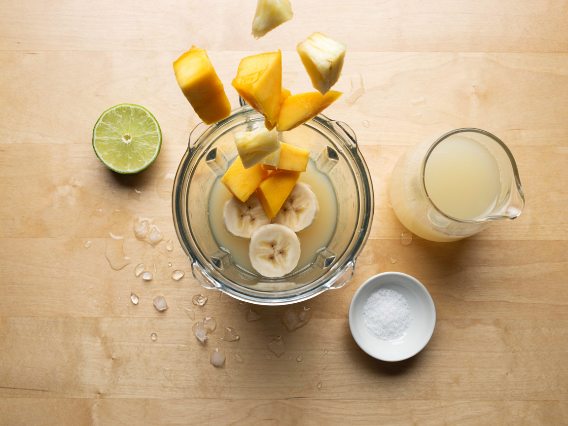 A blender jug on a light kitchen counter from above, coconut water and sliced banana in the bottom, frozen mango chunks and frozen pineapple being added on top, a halved lime, a small dish of salt and an open carton-free glass pitcher of coconut water beside it, bright kitchen light, overhead angle