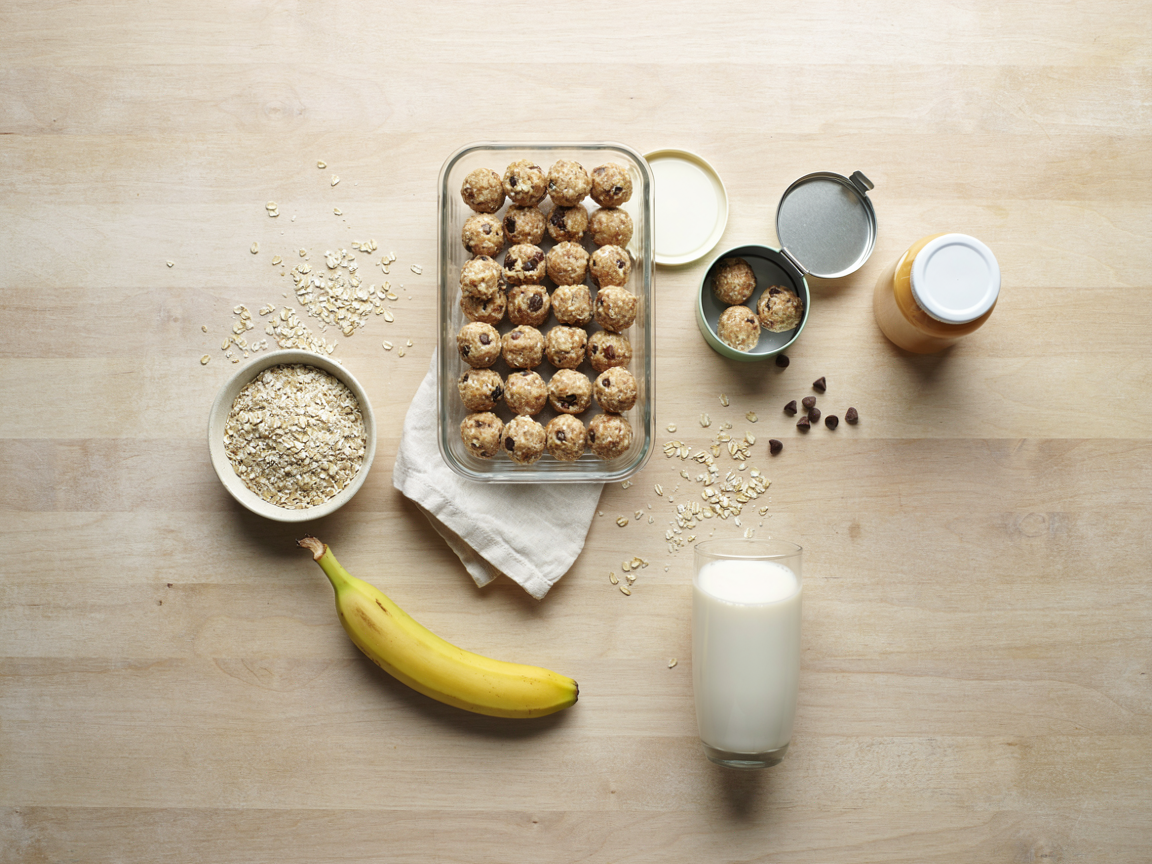 Weekly meal prep from above: a rectangular glass container filled with rows of oat protein balls with chocolate chips, two balls in a small open lunchbox tin, a jar of natural peanut butter, a bowl of rolled oats, a banana and a glass of milk on a light wooden table, overhead flat lay, bright natural light