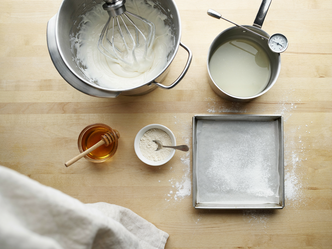 A stand mixer bowl from above with thick, glossy, nearly white marshmallow mixture clinging to the raised whisk in ribbons, a deep saucepan with a clip-on candy thermometer beside it, a small jar of honey, a bowl of powdered gelatin and a parchment-lined square pan dusted with white powder on a light counter, bright kitchen light