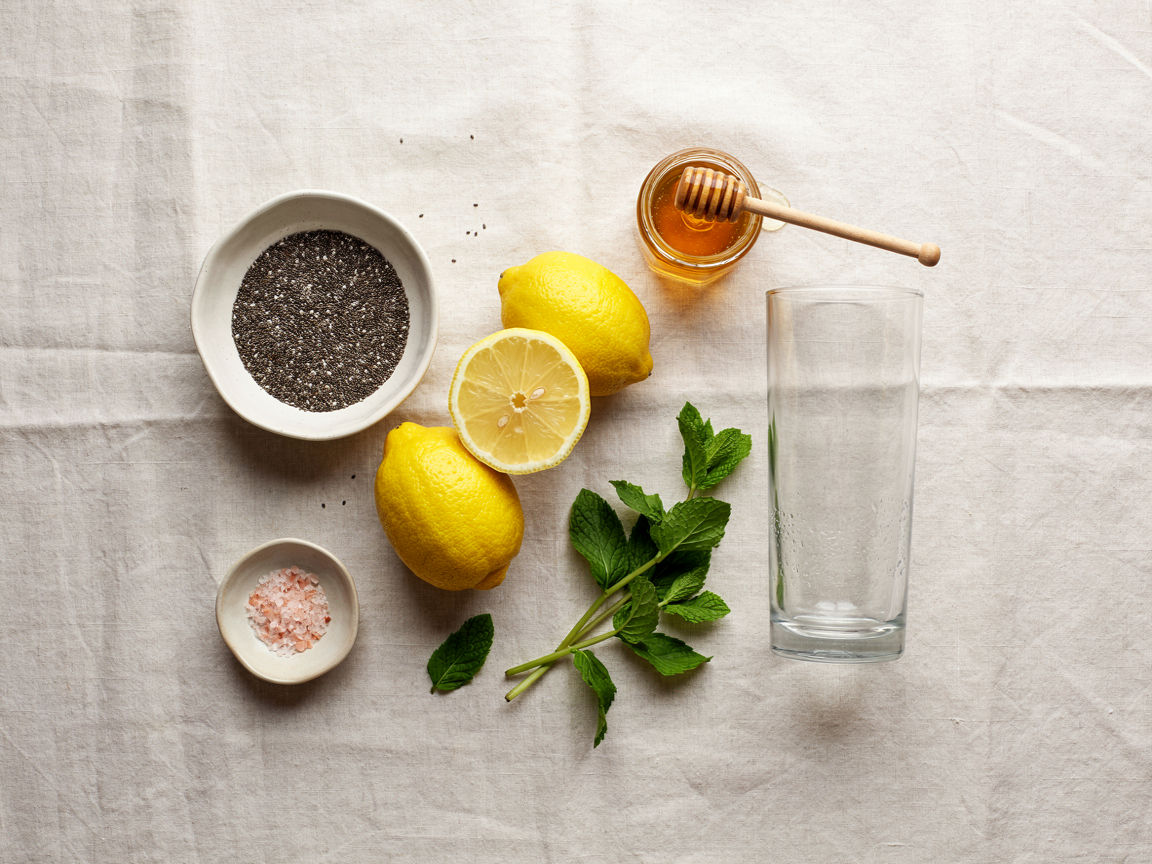 Ingredients for chia lemon water: a bowl of chia seeds, whole and halved lemons, fresh mint, pink salt, honey and an empty glass